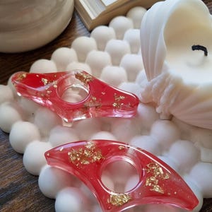 May include: Two red resin page holders with embedded gold leaf flakes. Each holder features a central circular opening and a pointed, angular shape. The items are displayed on a white, textured, bubble-patterned square tray.
