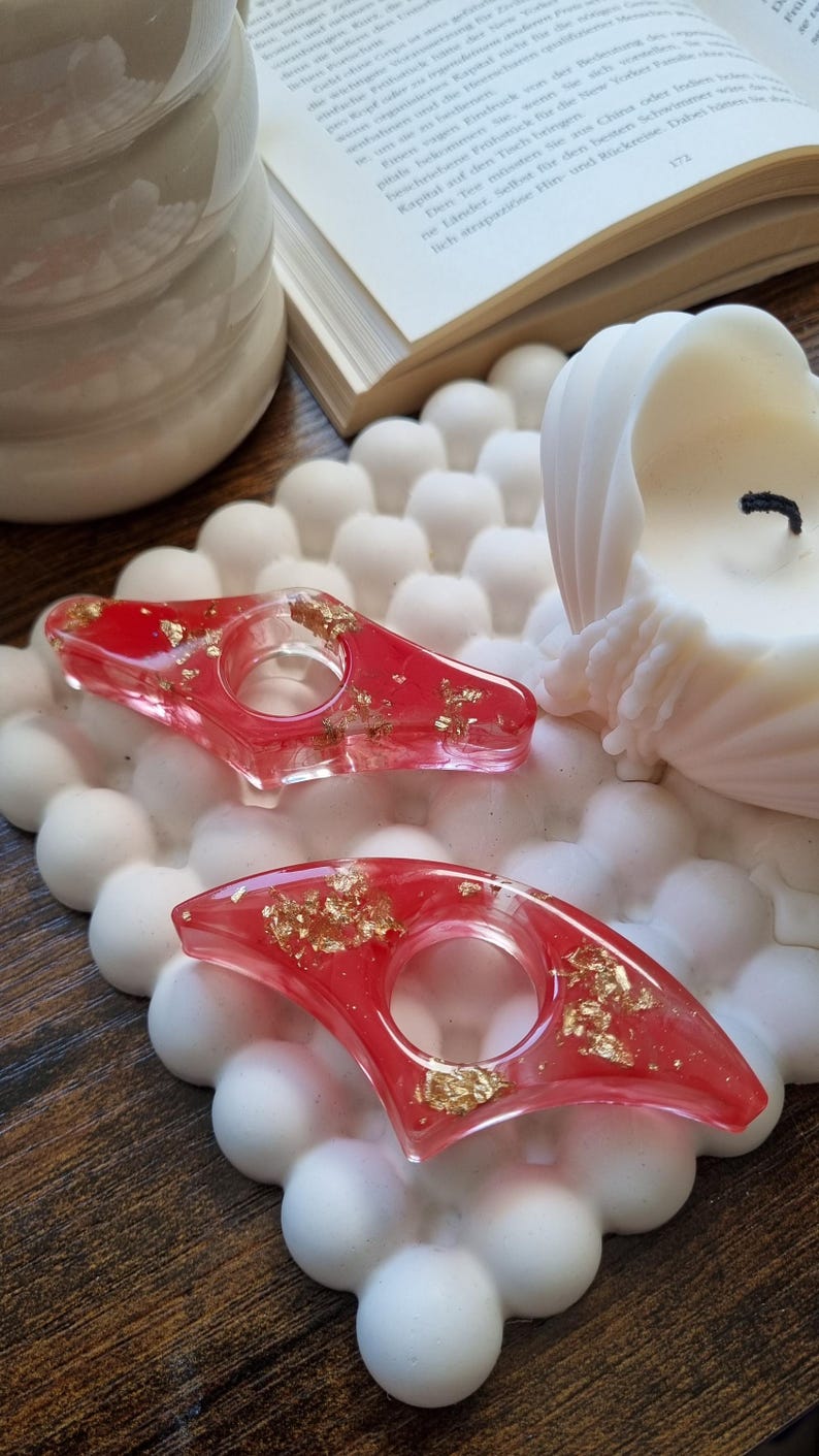 May include: Two red resin page holders with embedded gold leaf flakes. Each holder features a central circular opening and a pointed, angular shape. The items are displayed on a white, textured, bubble-patterned square tray.