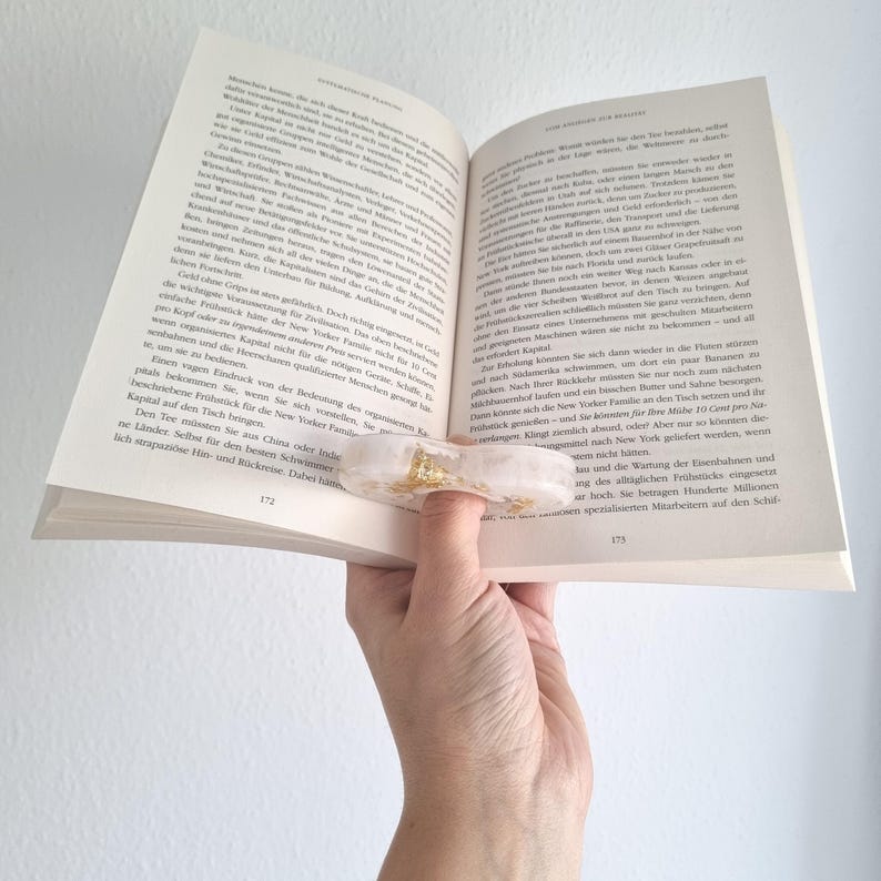 May include: A translucent, off-white resin book page holder with embedded gold leaf flakes. The oval-shaped accessory features a central circular opening designed to fit over a thumb, allowing for hands-free reading of an open book.