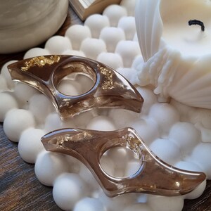 May include: Two crescent-shaped resin page holders in a translucent brown tint, featuring embedded gold-colored metallic flakes. Each holder has a central circular opening designed to fit over a finger for holding open book pages.