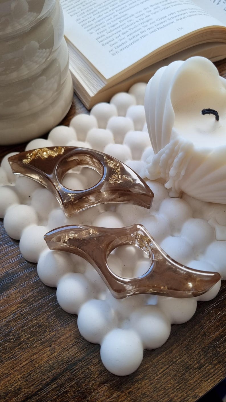 May include: Two crescent-shaped resin page holders in a translucent brown tint, featuring embedded gold-colored metallic flakes. Each holder has a central circular opening designed to fit over a finger for holding open book pages.