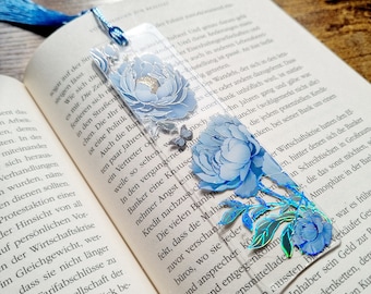 May include: A rectangular clear acrylic bookmark featuring blue floral illustrations with gold and iridescent foil accents. The bookmark includes a small butterfly graphic and a blue silk tassel attached to the top through a circular hole.