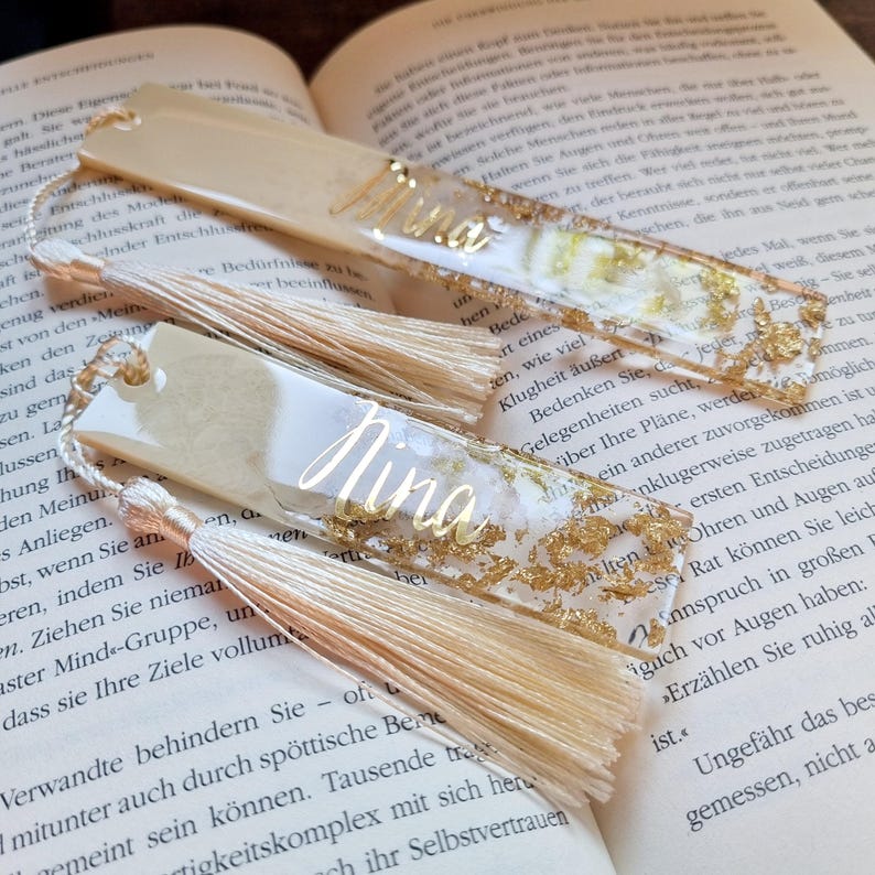 May include: Two rectangular resin bookmarks featuring a cream and white marbled design with embedded gold leaf flakes. Each bookmark displays the name Nina in gold cursive script and is finished with a long, light beige silk tassel attached.
