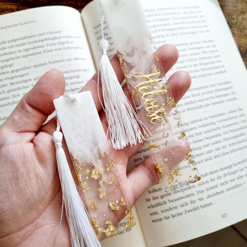 May include: Two rectangular resin bookmarks featuring a white opaque top section and a clear bottom section filled with gold leaf flakes. Each bookmark includes a white tassel attached to the top and one displays the name Heloise in gold script.