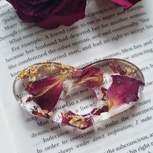 Könnte beinhalten: Ein transparentes herzförmiges Harz-Ornament mit getrockneten Rosenblättern und goldenen Blatt-Akzenten. Das Ornament liegt auf einem Buch mit dem Text "Um es zu überwinden, mache die Aussage "Ich bin immer unter direkter Inspiration; ich mache schnell."