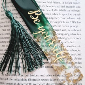 May include: A rectangular resin bookmark featuring a gradient from dark forest green to clear, embedded with gold leaf flakes. The word "Believe" is written in gold cursive script across the center. A dark green tassel is attached to the top.