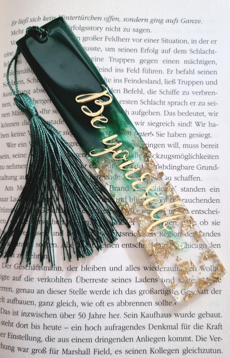 May include: A rectangular resin bookmark featuring a gradient from dark forest green to clear, embedded with gold leaf flakes. The word "Believe" is written in gold cursive script across the center. A dark green tassel is attached to the top.