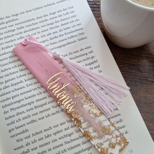 May include: A rectangular resin bookmark featuring a pink marbled top section and a clear bottom section filled with gold leaf flakes. The name Antonia is written in gold script across the center, and a light pink tassel is attached to the top.