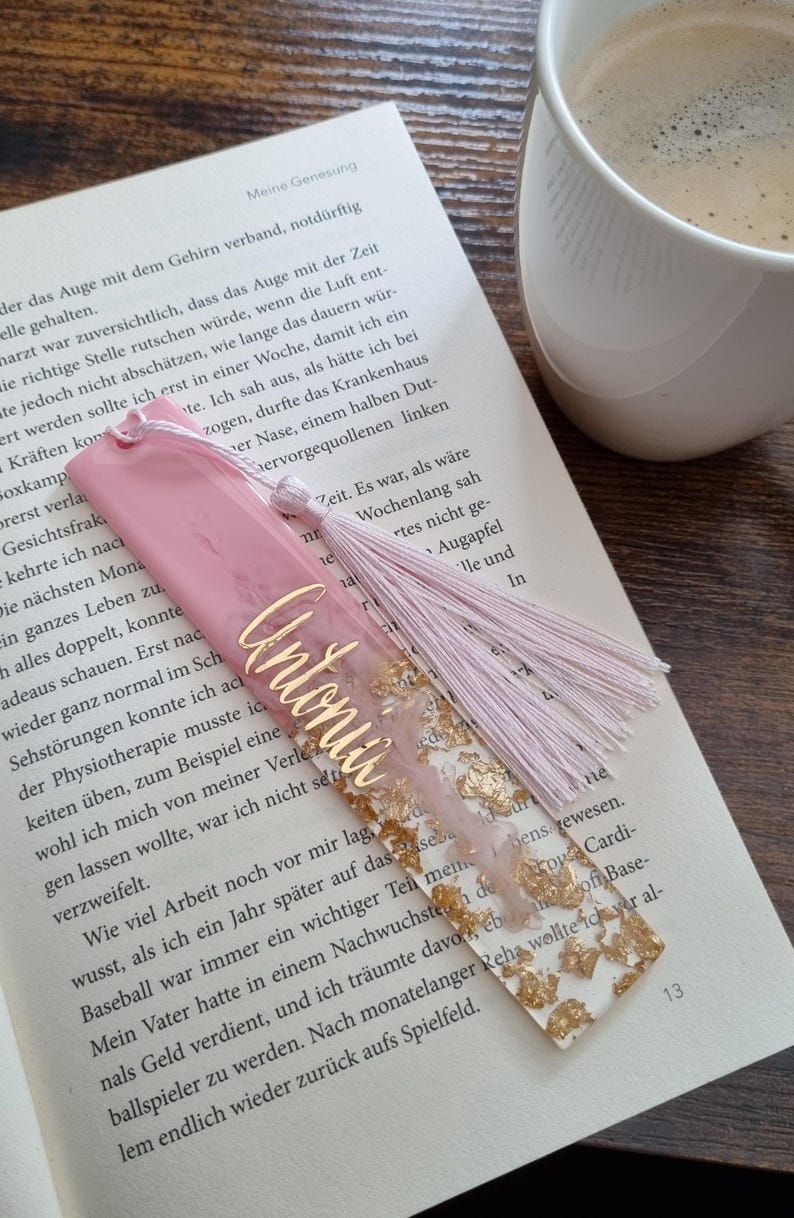May include: A rectangular resin bookmark featuring a pink marbled top section and a clear bottom section filled with gold leaf flakes. The name Antonia is written in gold script across the center, and a light pink tassel is attached to the top.