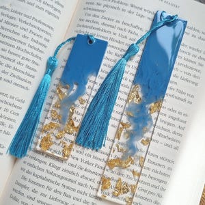 May include: Two rectangular resin bookmarks featuring a clear base with embedded gold leaf flakes and a swirling blue pigment design at the top. Each bookmark includes a hole at the top with an attached bright blue silk tassel for decoration.