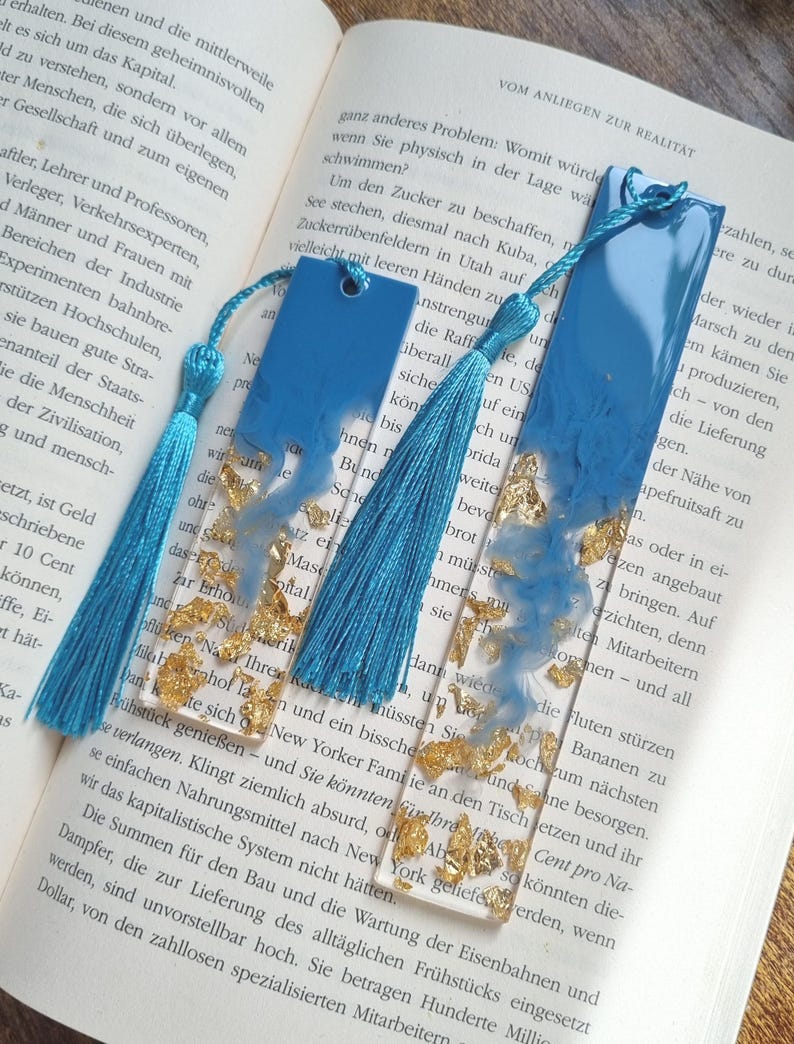May include: Two rectangular resin bookmarks featuring a clear base with embedded gold leaf flakes and a swirling blue pigment design at the top. Each bookmark includes a hole at the top with an attached bright blue silk tassel for decoration.