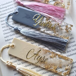 May include: Three rectangular resin bookmarks with tassels. The top bookmark is pink with gold flakes and the name Oma. The middle bookmark is gray with gold flakes and the name Yunus. The bottom bookmark is cream with gold flakes and the name Ayse.