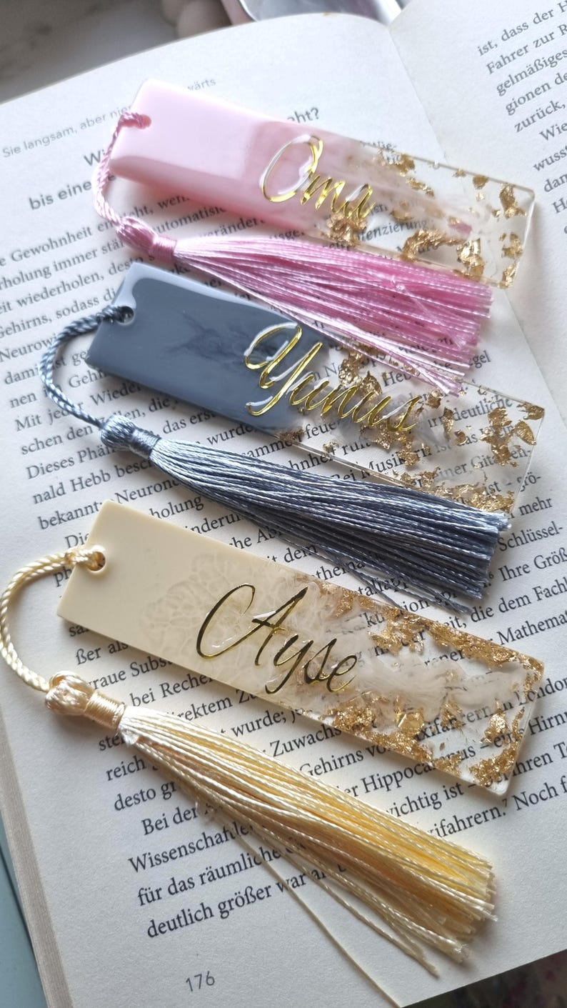 May include: Three rectangular resin bookmarks with tassels. The top bookmark is pink with gold flakes and the name Oma. The middle bookmark is gray with gold flakes and the name Yunus. The bottom bookmark is cream with gold flakes and the name Ayse.