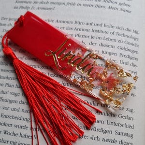May include: A rectangular resin bookmark featuring a vibrant red base with embedded gold leaf flakes and a pink floral design. The name Linda is written in gold cursive script on the surface. A long, bright red fabric tassel is attached to the top.