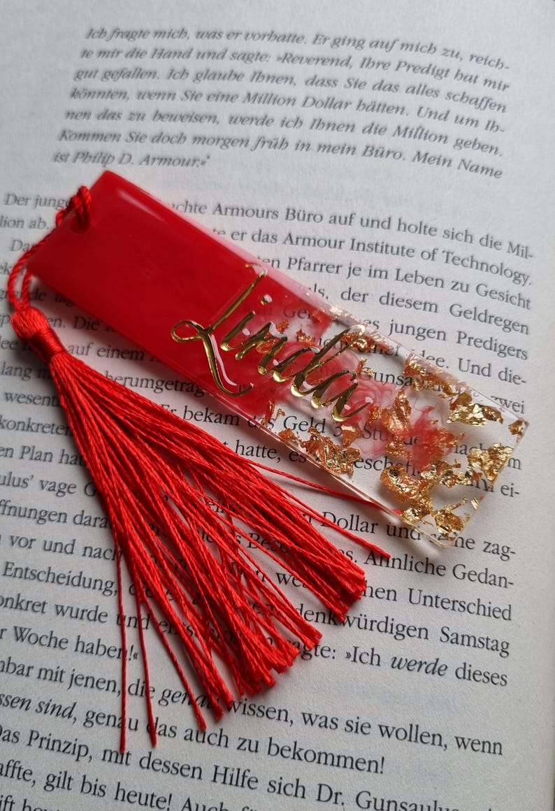 May include: A rectangular resin bookmark featuring a vibrant red base with embedded gold leaf flakes and a pink floral design. The name Linda is written in gold cursive script on the surface. A long, bright red fabric tassel is attached to the top.