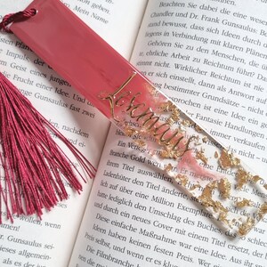 May include: A rectangular bookmark featuring a solid dark pink top section and a clear bottom section filled with gold leaf flakes. The word Lesemaus is written in gold cursive script across the clear portion. A dark pink tassel is attached to the top.
