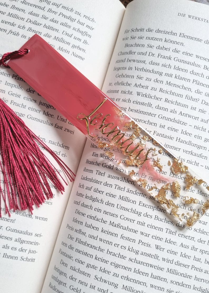 May include: A rectangular bookmark featuring a solid dark pink top section and a clear bottom section filled with gold leaf flakes. The word Lesemaus is written in gold cursive script across the clear portion. A dark pink tassel is attached to the top.