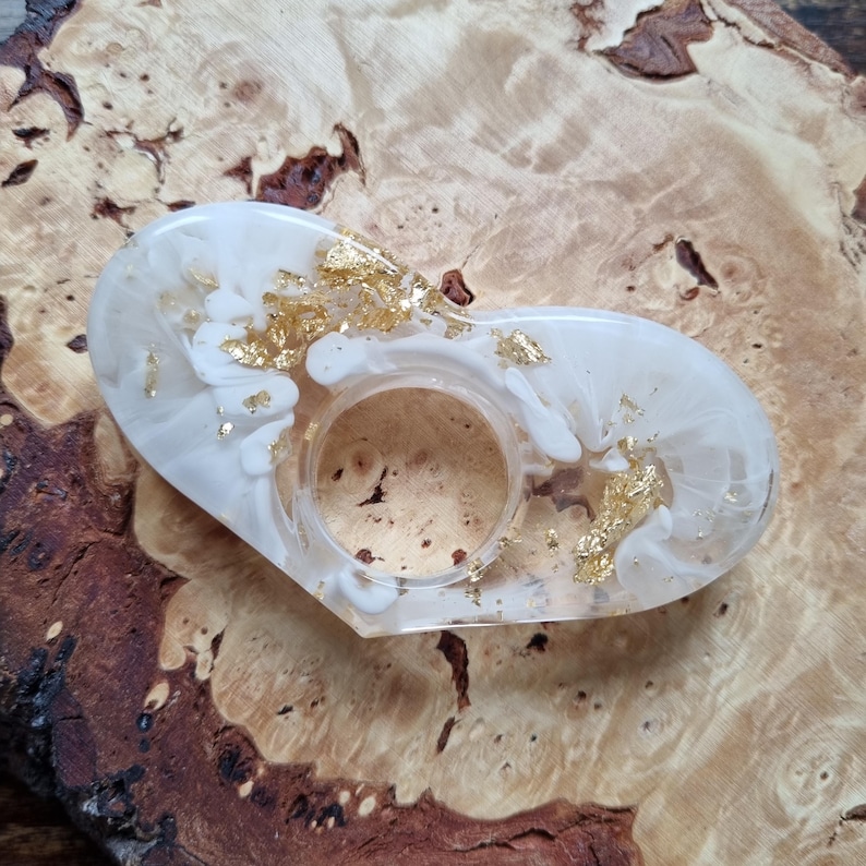 May include: A heart-shaped resin ring holder featuring a white marbled pattern with embedded gold leaf flakes. The object has a smooth, glossy finish and a central circular opening designed to hold a single ring securely in place.