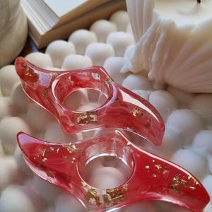 May include: Two resin page holders with a translucent red marbled design and embedded gold leaf flakes. Each holder features a central circular opening designed to slide over a thumb for holding open book pages while reading.