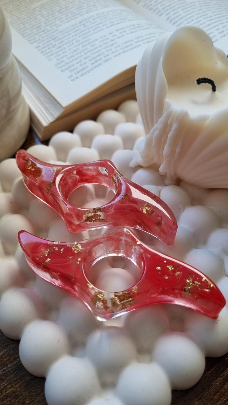 May include: Two resin page holders with a translucent red marbled design and embedded gold leaf flakes. Each holder features a central circular opening designed to slide over a thumb for holding open book pages while reading.