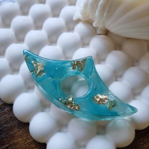 May include: A crescent-shaped teal resin ring featuring embedded gold leaf flakes. The ring has a smooth, translucent finish with a central circular opening, showcasing a marbled color effect throughout the material.