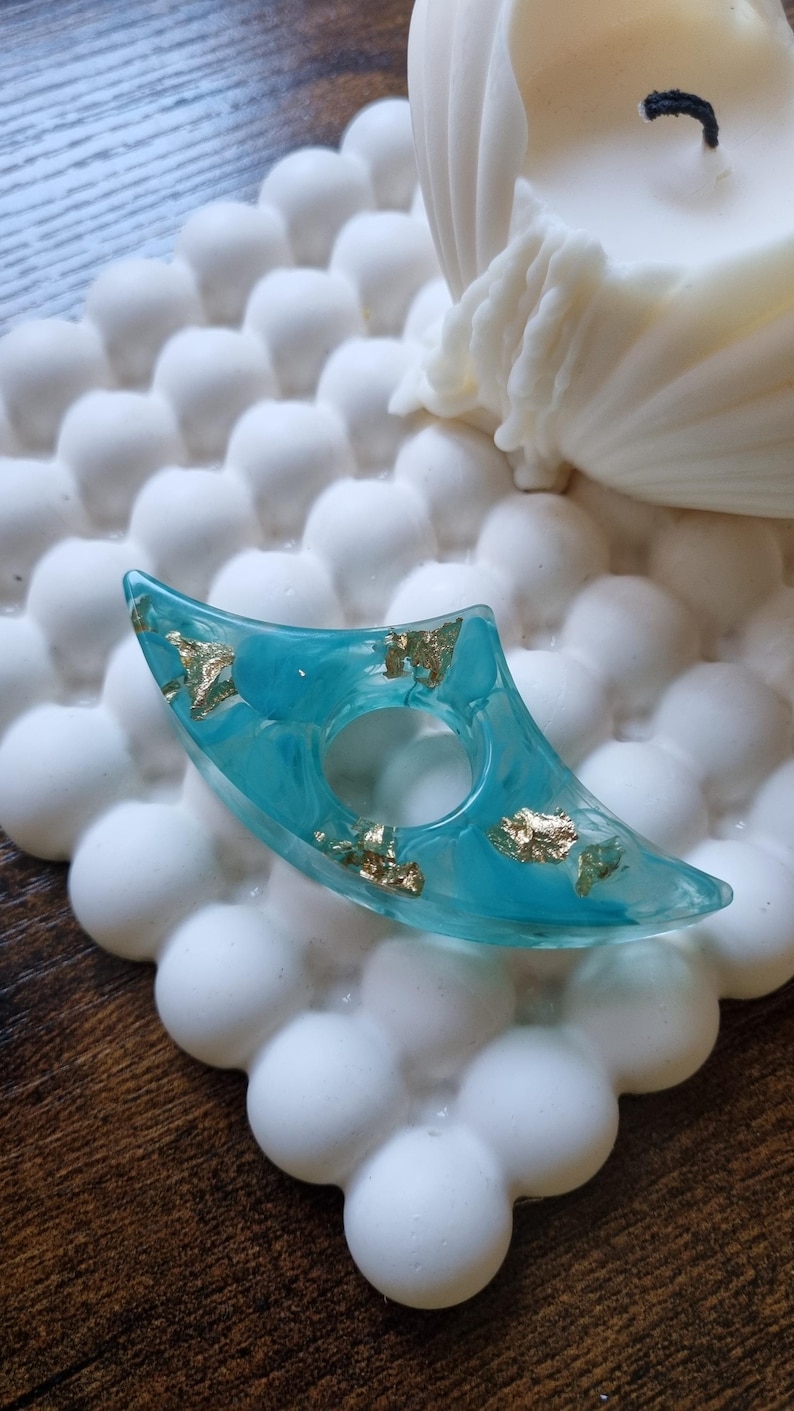 May include: A crescent-shaped teal resin ring featuring embedded gold leaf flakes. The ring has a smooth, translucent finish with a central circular opening, showcasing a marbled color effect throughout the material.