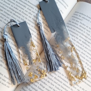 May include: Two rectangular resin bookmarks featuring a blend of solid gray and clear sections with embedded gold leaf flakes. Each bookmark is topped with a hole and a matching gray silk tassel, resting on the open pages of a printed book.