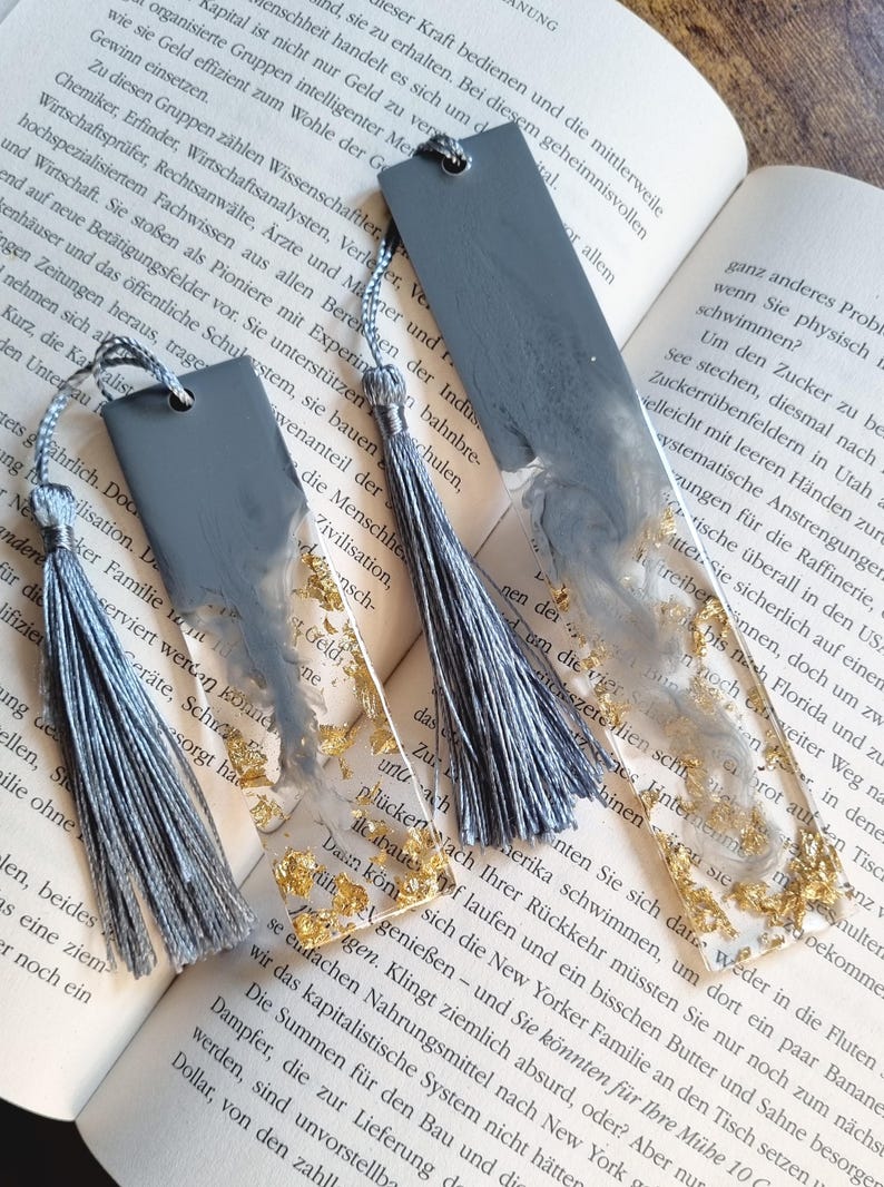 May include: Two rectangular resin bookmarks featuring a blend of solid gray and clear sections with embedded gold leaf flakes. Each bookmark is topped with a hole and a matching gray silk tassel, resting on the open pages of a printed book.
