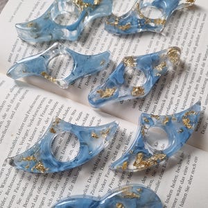 Könnte beinhalten: Ein Satz von fünf blauen und goldenen Lesezeichenringen aus Harz. Die Ringe sind halbmondförmig und haben ein Blattgold-Design.