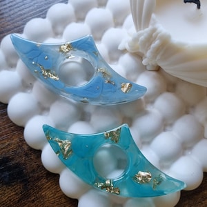May include: Two crescent-shaped resin rings featuring a smooth finish with embedded gold leaf flakes. One ring is a light sky blue, while the other is a vibrant turquoise. Both items have a central circular opening for a finger.