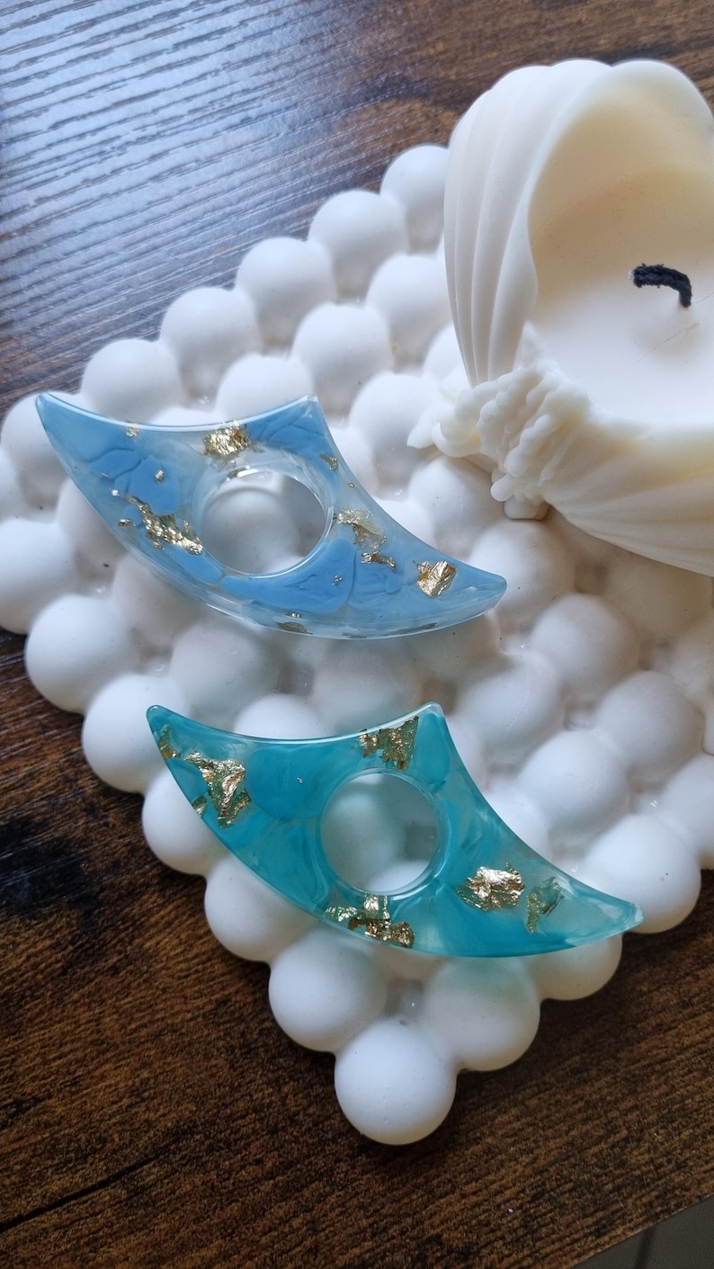 May include: Two crescent-shaped resin rings featuring a smooth finish with embedded gold leaf flakes. One ring is a light sky blue, while the other is a vibrant turquoise. Both items have a central circular opening for a finger.