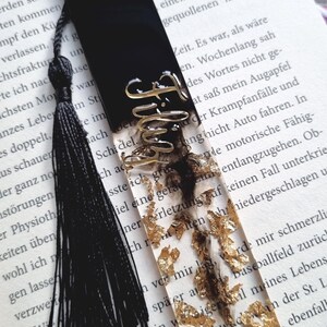 May include: A rectangular resin bookmark featuring a solid black top section and a clear bottom section filled with gold leaf flakes. The word Filthy is written in gold cursive script across the center. A black tassel is attached to the top.