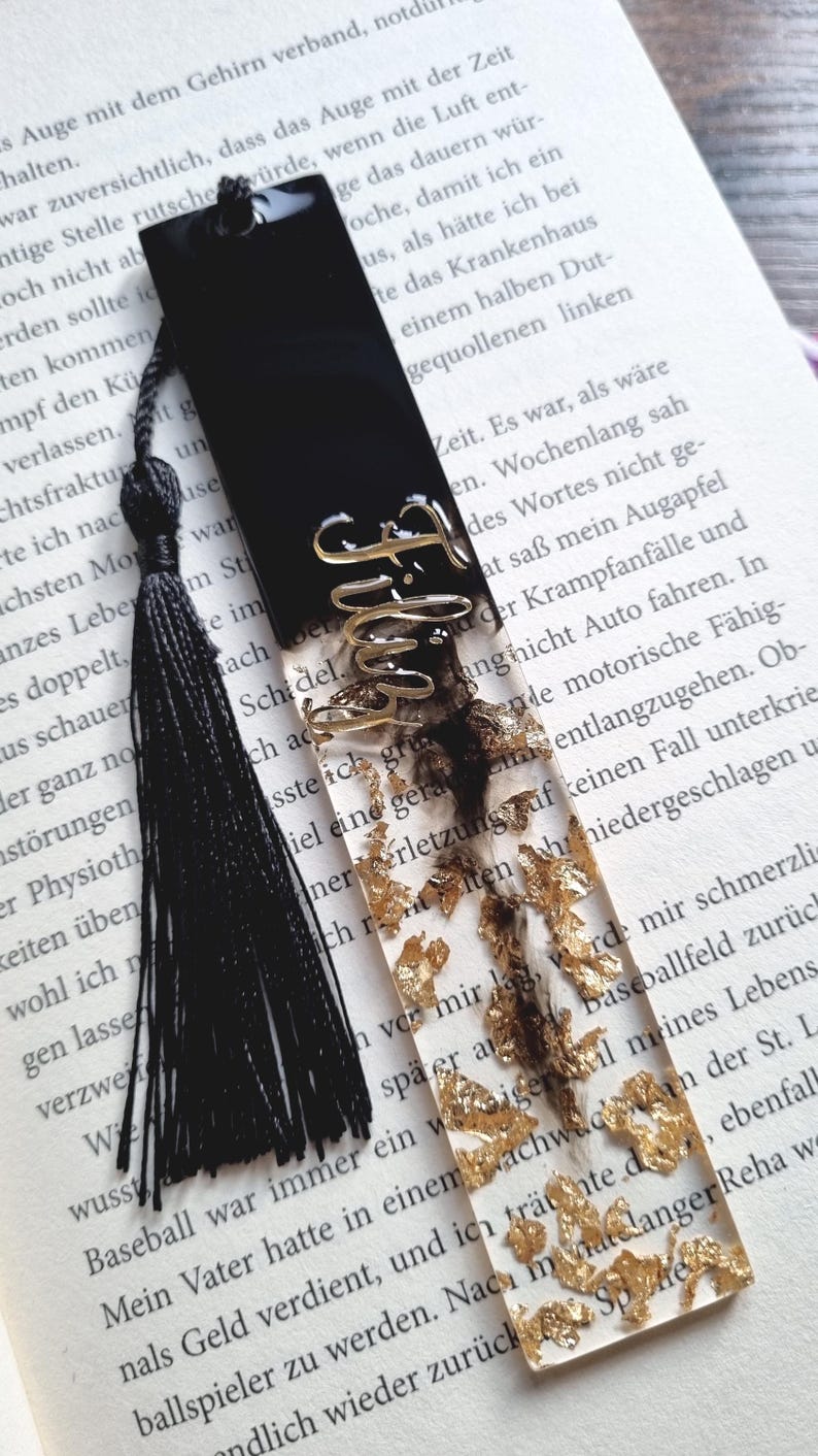 May include: A rectangular resin bookmark featuring a solid black top section and a clear bottom section filled with gold leaf flakes. The word Filthy is written in gold cursive script across the center. A black tassel is attached to the top.