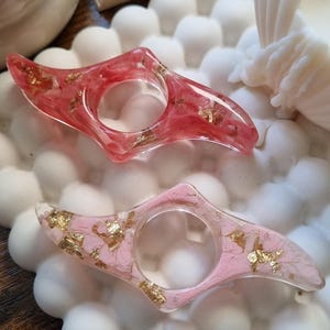 May include: Two resin finger rings with a pointed, elongated shape. The top ring is translucent red with embedded gold leaf flakes. The bottom ring is translucent light pink with embedded gold leaf flakes. Both rings feature a central circular opening.