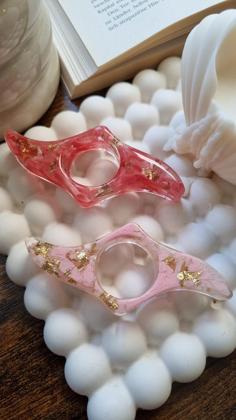 May include: Two resin finger rings with a pointed, elongated shape. The top ring is translucent red with embedded gold leaf flakes. The bottom ring is translucent light pink with embedded gold leaf flakes. Both rings feature a central circular opening.