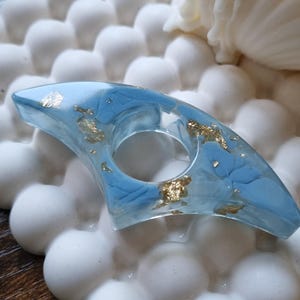 May include: A crescent-shaped ring made of clear resin with embedded light blue pigment swirls and scattered gold leaf flakes. The ring features a central circular opening and a smooth, polished finish throughout its entire surface.