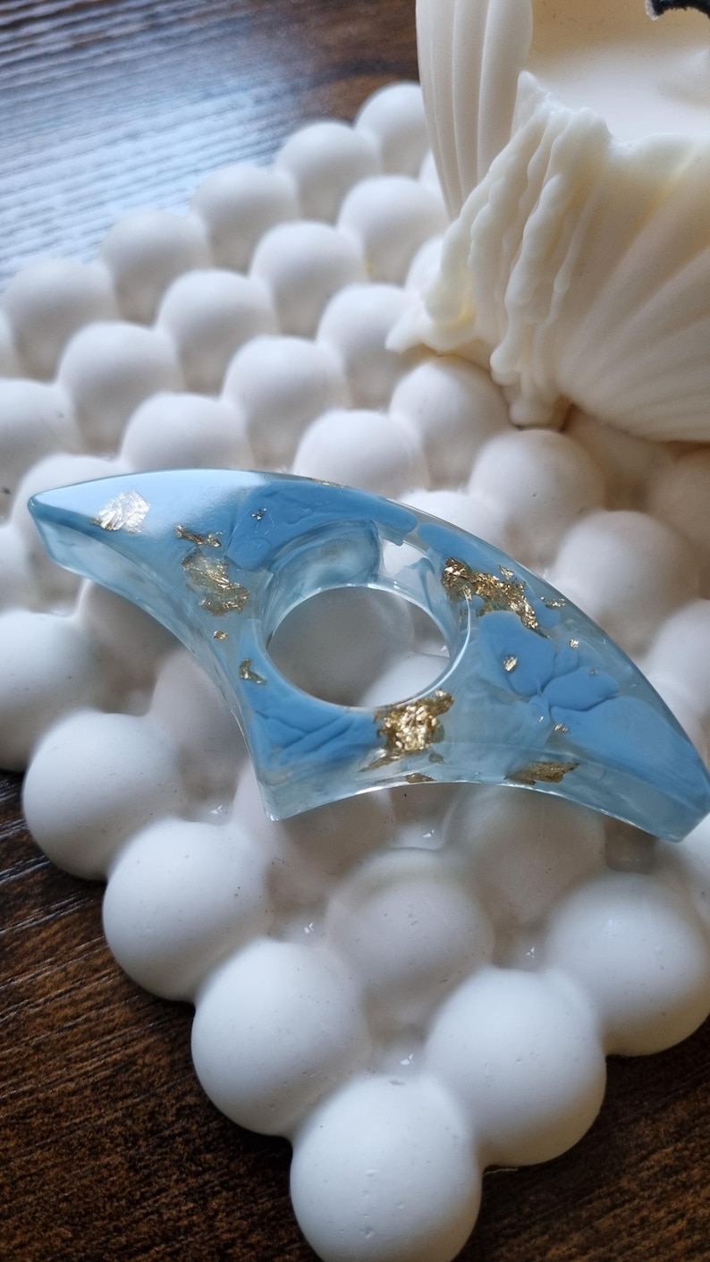 May include: A crescent-shaped ring made of clear resin with embedded light blue pigment swirls and scattered gold leaf flakes. The ring features a central circular opening and a smooth, polished finish throughout its entire surface.