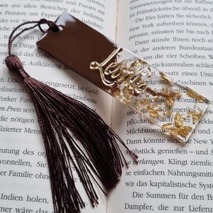 May include: A rectangular bookmark featuring a solid dark brown section and a clear section filled with gold leaf flakes. The name Leonie is written in gold script across the clear portion. A dark brown tassel is attached to the top hole.