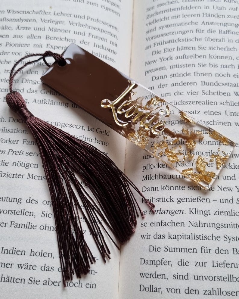 May include: A rectangular bookmark featuring a solid dark brown section and a clear section filled with gold leaf flakes. The name Leonie is written in gold script across the clear portion. A dark brown tassel is attached to the top hole.