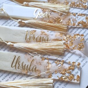 May include: A collection of five rectangular bookmarks made of translucent resin with embedded gold leaf flakes. Each bookmark features a personalized name in gold script lettering and is finished with a matching cream-colored silk tassel.