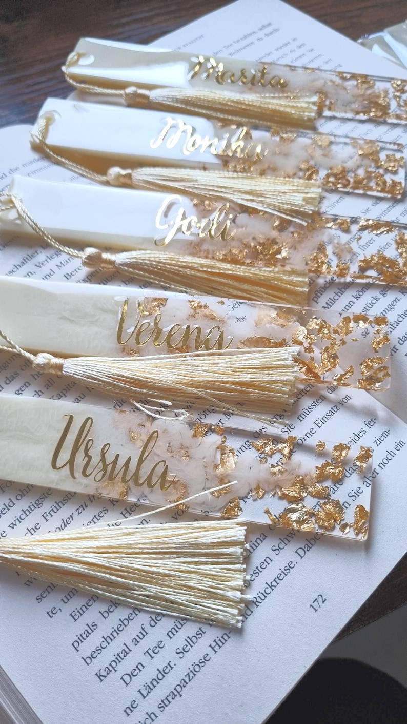 May include: A collection of five rectangular bookmarks made of translucent resin with embedded gold leaf flakes. Each bookmark features a personalized name in gold script lettering and is finished with a matching cream-colored silk tassel.