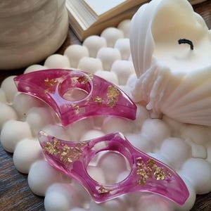 May include: Two crescent-shaped resin candle holders in a translucent magenta pink hue, featuring embedded gold leaf flakes throughout. Each piece has a circular opening in the center, designed to hold a taper candle securely.