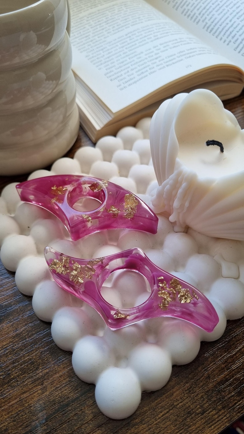 May include: Two crescent-shaped resin candle holders in a translucent magenta pink hue, featuring embedded gold leaf flakes throughout. Each piece has a circular opening in the center, designed to hold a taper candle securely.