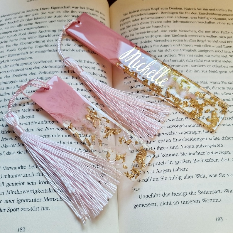 May include: Two rectangular resin bookmarks featuring a dusty rose and clear design with embedded gold leaf flakes. Each bookmark is finished with a light pink silk tassel attached through a small hole. One bookmark displays the name Michelle in white script.