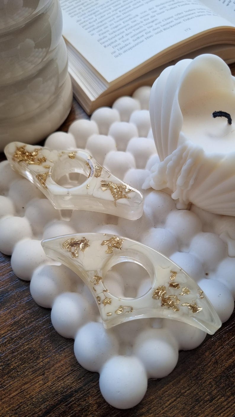 May include: Two crescent-shaped resin napkin rings featuring a translucent off-white base embedded with scattered gold leaf flakes. Each ring has a central circular opening and a smooth, polished finish, showcasing a distinct curved design.