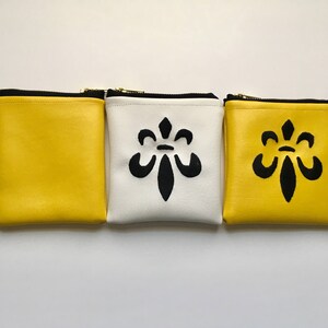 Puede incluir: Tres pequeñas bolsas con cremallera, una amarilla, una blanca y una amarilla. Cada bolsa tiene un diseño de flor de lis negra.