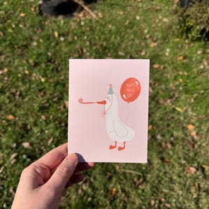 Pu&ograve; includere: Un biglietto d'auguri rosa con un'illustrazione di un'oca bianca con un cappello da festa e un palloncino rosso che dice "Happy Hatch Day".