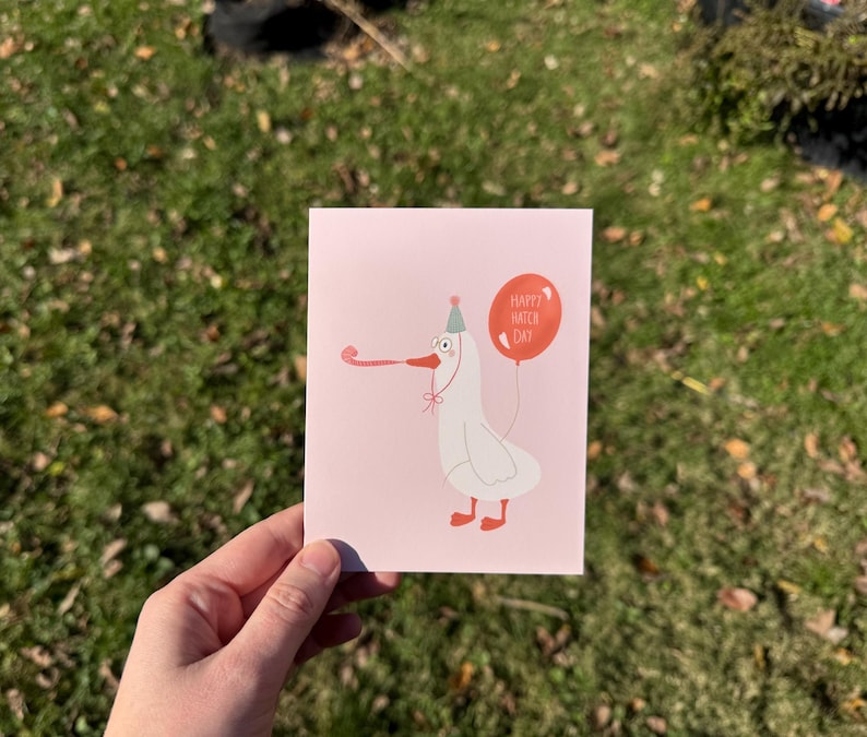 Pu&ograve; includere: Un biglietto d'auguri rosa con un'illustrazione di un'oca bianca con un cappello da festa e un palloncino rosso che dice "Happy Hatch Day".