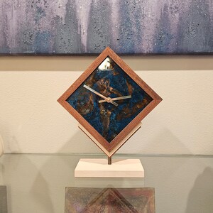 May include: A diamond-shaped wooden clock with a blue and brown resin face. The clock has gold-colored hands and is mounted on a light wood stand. The clock is displayed on a glass surface.