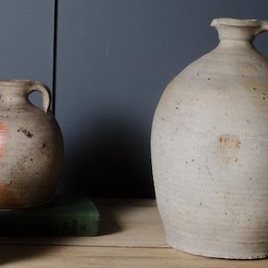 May include: Three ceramic jugs with rounded bodies and single handles. The largest jug is off-white with a speckled texture. Two smaller jugs feature a mix of beige and reddish-brown tones with visible surface wear and earthy, weathered finishes.